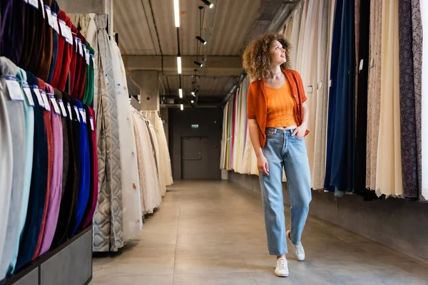 Full length of curly woman holding hand in pocket of jeans and looking at curtains in textile shop — Stock Photo
