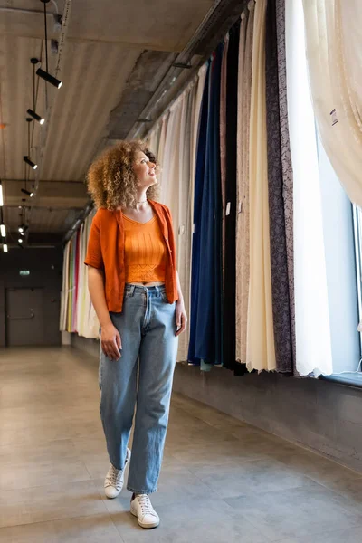 Full length of pleased curly woman in jeans looking at colorful curtains in textile shop — Stock Photo