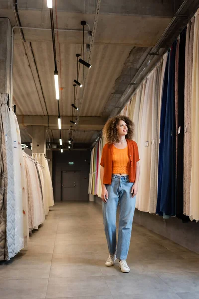 Full length of curly woman in jeans walking along assortment of curtains and fabric in textile shop — Stock Photo