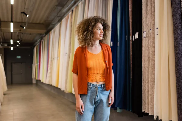 Smiling woman with curly hair looking at multicolored curtains in textile shop — Stock Photo