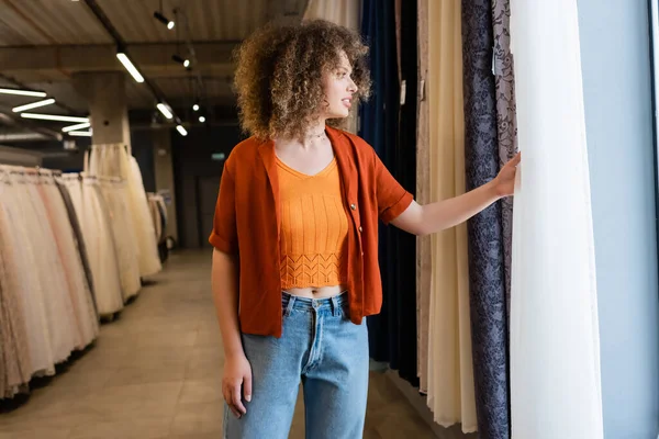 Seitenansicht einer lächelnden, lockigen Frau mit Blick auf Vorhänge in einem Textilgeschäft — Stockfoto