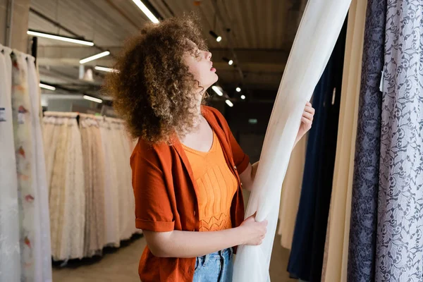 Vista lateral da jovem mulher encaracolada escolhendo cortinas na loja têxtil — Fotografia de Stock