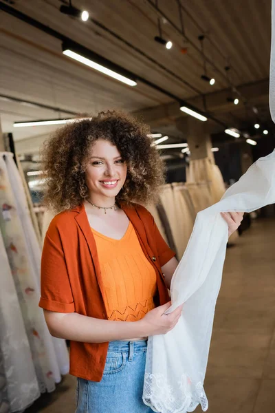 Curly woman smiling at camera while choosing curtains in textile shop — Stock Photo