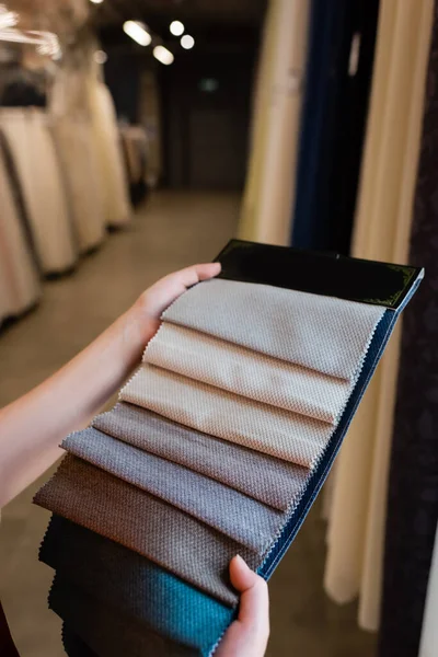 Vista recortada de una mujer joven sosteniendo muestras de tela en una tienda textil borrosa - foto de stock