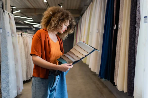 Side view of curly client holding fabric samples in blurred textile shop — Stock Photo