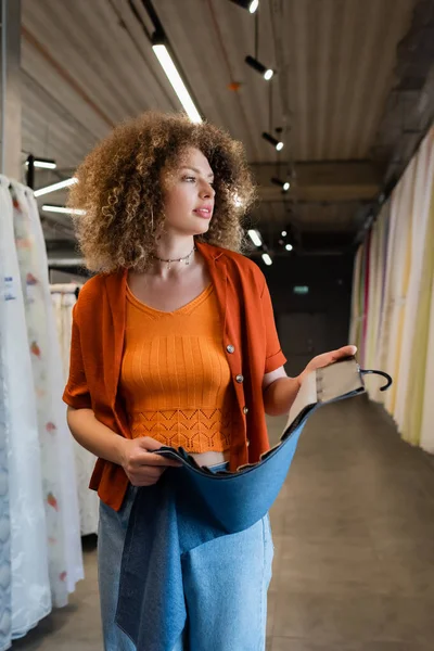 Молодая кудрявая женщина держит образцы ткани и смотрит прочь в текстильном магазине — стоковое фото