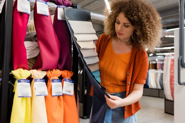 Femme bouclée regardant des échantillons de tissu près de tissu dans un magasin de textile — Photo de stock