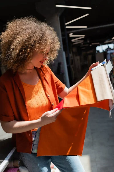 Curly customer choosing colorful cloth while holding samples in textile shop — Stock Photo