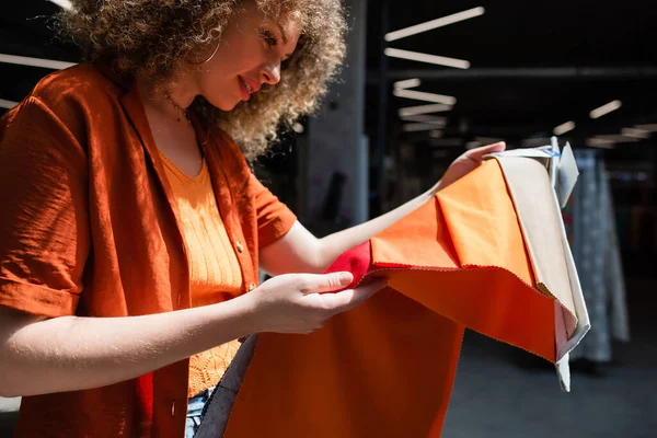 Heureuse femme bouclée tenant des échantillons de textiles colorés dans un magasin flou — Photo de stock
