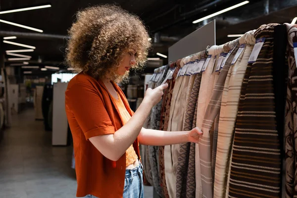 Side view of curly buyer taking photo on smartphone while choosing fabric in textile shop — Stock Photo