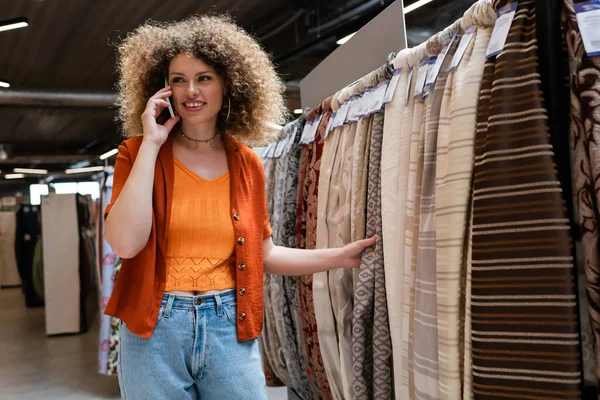 Mujer sonriente hablando por teléfono celular y mirando hacia otro lado cerca de la tela en la tienda textil - foto de stock