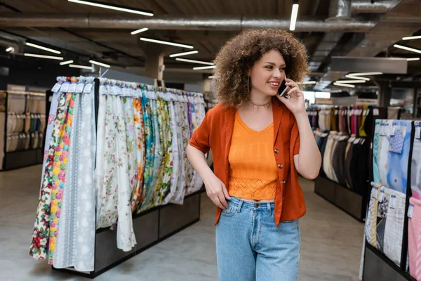 Cliente alegre hablando en el teléfono inteligente en la tienda textil borrosa - foto de stock