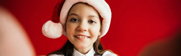 Портрет веселой девушки в шляпе Санты, смотрящей на камеру, изолированную на красном, баннер — стоковое фото