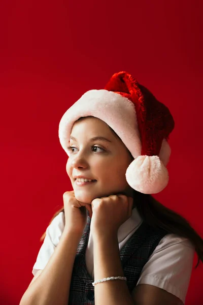 Chica sonriente y soñadora en sombrero de santa cogida de la mano cerca de la barbilla y mirando hacia otro lado aislado en rojo - foto de stock