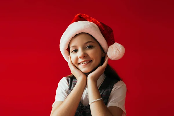 Smiling girl in santa hat and beaded bracelet holding hands near face isolated on red — Stock Photo