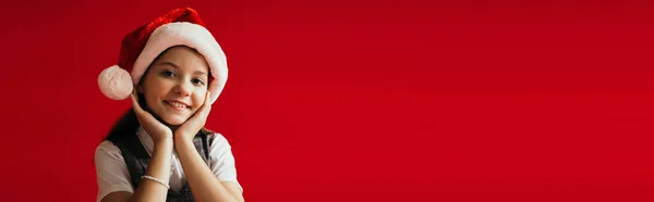 Gioiosa ragazza in cappello di Babbo Natale sorridente alla fotocamera mentre si tiene le mani vicino al viso isolato sul rosso, banner — Foto stock