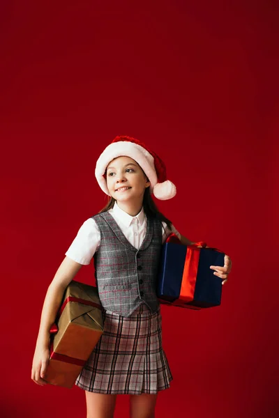Gaie fille dans santa chapeau et vêtements à la mode tenant boîtes-cadeaux et regardant loin isolé sur rouge — Photo de stock