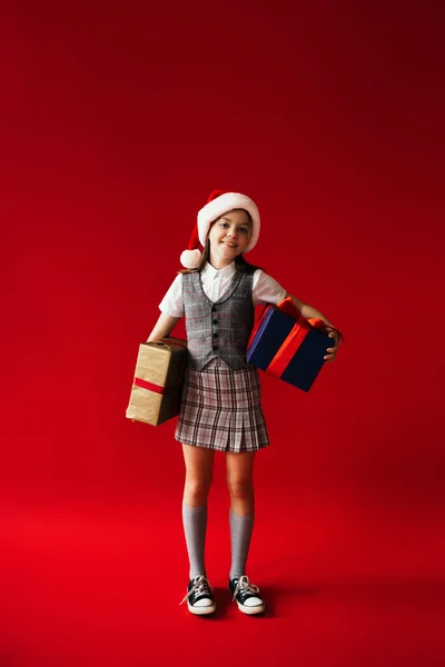 Comprimento total da menina na moda e feliz em santa chapéu e roupas xadrez segurando caixas de presente no fundo vermelho — Fotografia de Stock