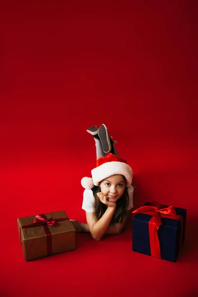 Chica feliz en sombrero de santa mintiendo cerca de regalos de año nuevo y mirando a la cámara en el fondo rojo - foto de stock
