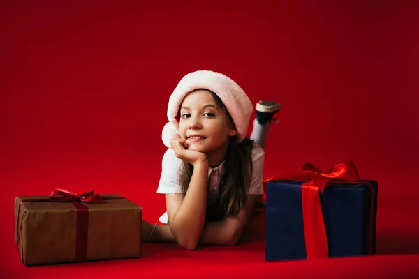Allegro preteen ragazza in cappello di Babbo Natale guardando la fotocamera mentre si trova vicino a regali di Natale su sfondo rosso — Foto stock