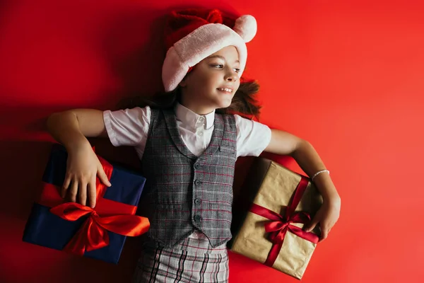 Draufsicht des fröhlichen Mädchens mit Weihnachtsmütze und karierter Weste, das in der Nähe von Geschenken auf rotem Hintergrund liegt — Stockfoto