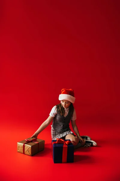 Повна довжина дівчини в капелюсі Санта і капелюсі, що сидить біля подарункових коробок на червоному тлі з місцем для тексту — Stock Photo