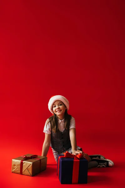 Cheerful girl in santa hat sitting near new year presents on red background with copy space — Stock Photo