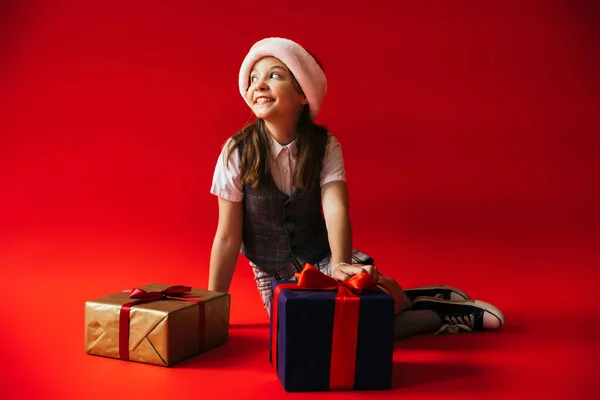 Joyful child in plaid vest and santa hat sitting near gift boxes and looking away on red background — Stock Photo