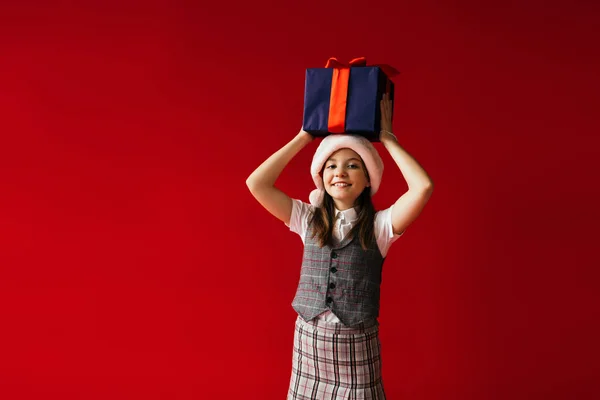 Cheerful girl in plaid clothes and santa hat holding Christmas presents above head isolated on red — Stock Photo