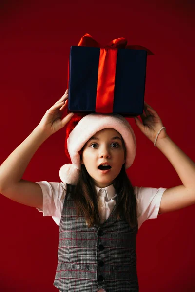 Amazed girl in santa hat standing with open mouth and gift box above head isolated on red — Stock Photo