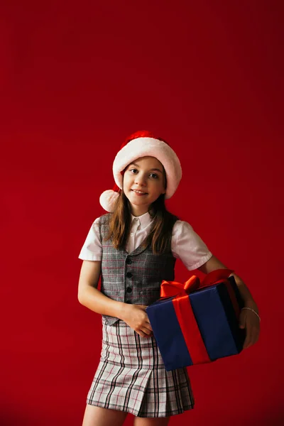 Smiling child in santa hat and plaid skirt standing with Christmas present isolated on red — Stock Photo