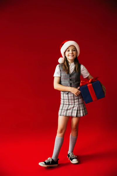 Volle Länge des Mädchens in Gummischuhen und kariertem Rock trägt Weihnachtsmann Hut und hält blaue Geschenkbox auf rotem Hintergrund — Stockfoto