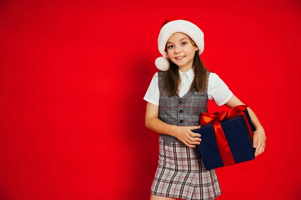 Preteen girl in stylish checkered clothes and santa hat holding Christmas present isolated on red — Stock Photo