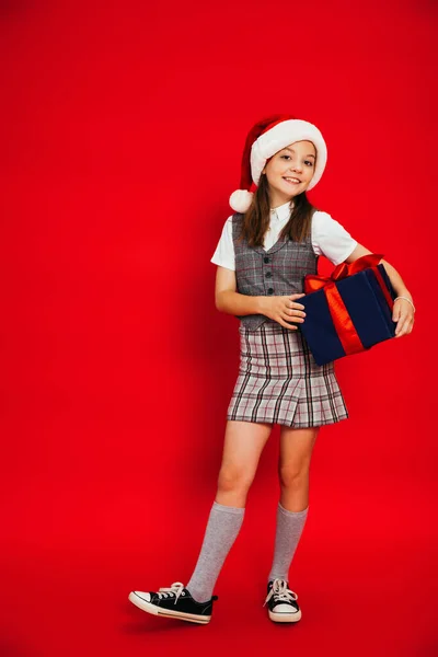 Comprimento total da criança alegre na saia xadrez e chapéu de santa segurando caixa de presente azul no fundo vermelho — Fotografia de Stock