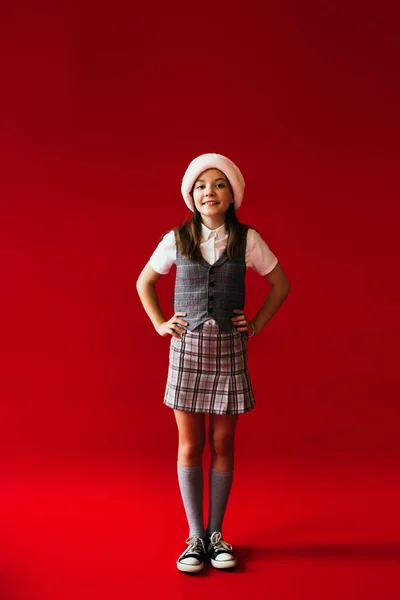 Volle Länge des Mädchens in kariertem Rock und Weihnachtsmütze posiert mit den Händen auf den Hüften auf rotem Hintergrund — Stockfoto