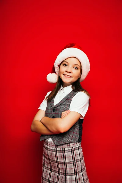 Smiling girl in santa hat and plaid vest standing with crossed arms isolated on red — Stock Photo