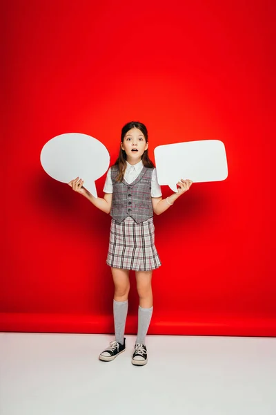 Volle Länge des Mädchens in kariertem Rock und Gummischuhen mit leeren Sprechblasen auf rotem Hintergrund — Stockfoto