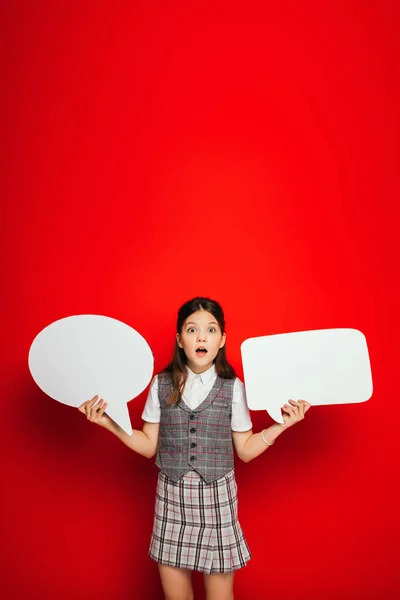 Amazed girl with open mouth holding white speech bubbles isolated on red — Stock Photo