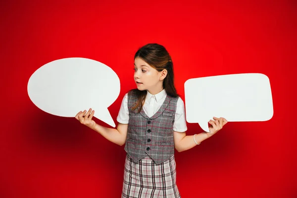 Girl in plaid skirt and vest holding white and empty speech bubbles isolated on red — Stock Photo