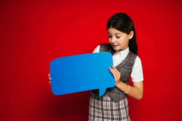 Smiling preteen girl in plaid skirt looking at blue speech bubble isolated on red — Stock Photo