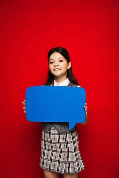 Joyful child in checkered skirt showing blue speech bubble at camera isolated on red — Stock Photo