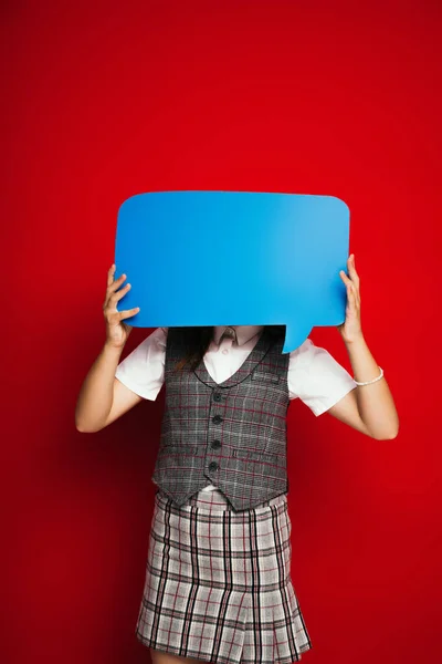 Preteen child in checkered vest and skirt obscuring face with blue speech bubble isolated on red — Stock Photo