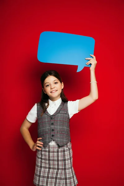 Preteen girl in plaid vest standing with hand on hip and holding blue speech bubble in raised hand isolated on red — Stock Photo