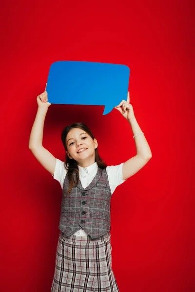 Menina pré-adolescente alegre em colete quadriculado mostrando bolha de fala azul isolado no vermelho — Fotografia de Stock