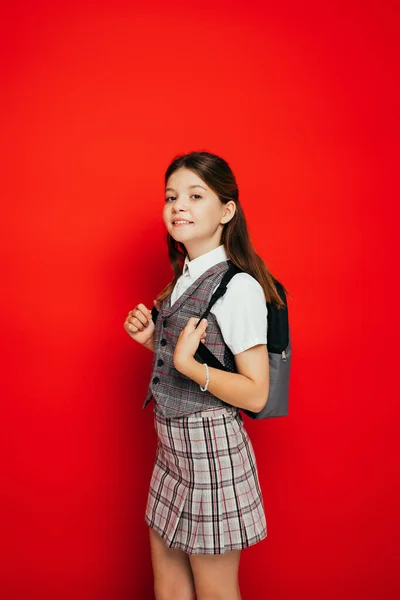 Chica positiva en chaleco a cuadros sonriendo a la cámara mientras está de pie con la mochila aislada en rojo, pancarta - foto de stock
