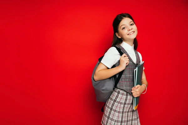 Elegante colegiala con copybooks y mochila sonriendo a la cámara aislada en rojo, pancarta - foto de stock