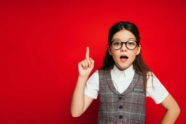 Astonished girl in eyeglasses showing idea gesture and looking at camera isolated on red — Stock Photo