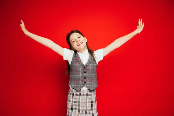 Веселая девушка в стильной клетчатой одежде стоит с протянутыми руками, изолированными на красном, баннер — стоковое фото