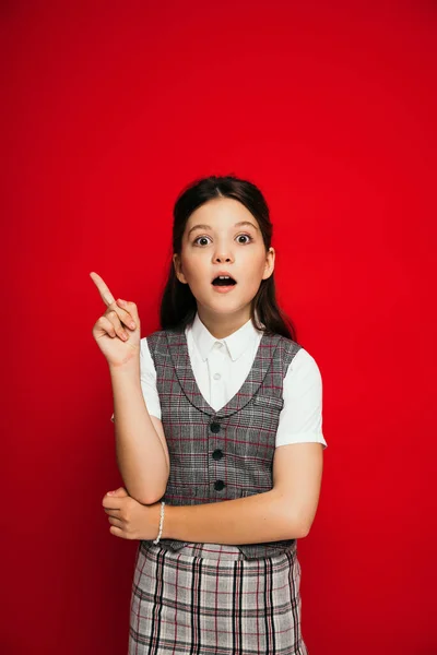 Astonished brunette girl in plaid vest looking at camera and pointing with finger isolated on red, banner — Stock Photo
