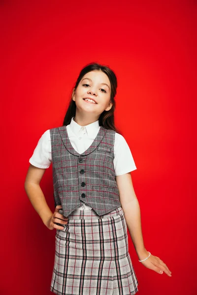 Cheerful brunette girl in plaid vest and skirt posing with hand on hip isolated on red, banner — Stock Photo